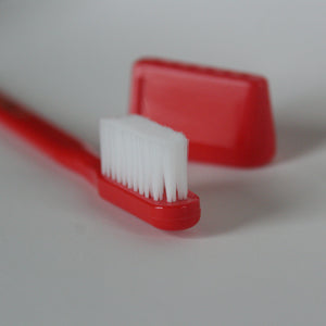 A toothbrush with even bristles and a red handle, with its triangular cap in the background.